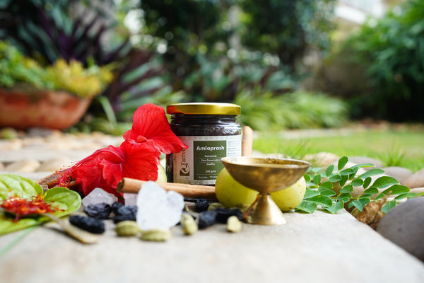 Tattved homemade Amlaprash jar, 200 gms, zero preservative, surrounded by amla, cinnamon, cardamom, hibiscus, and a brass bowl in a garden setting