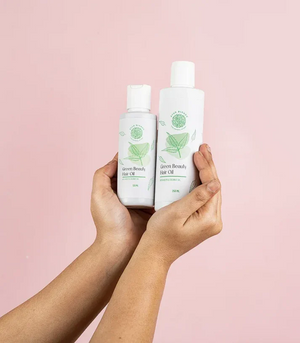 Hands holding two sizes of Ayur Blends Green Beauty Hair Oil, 120ml and 250ml, made with neem and coconut oil