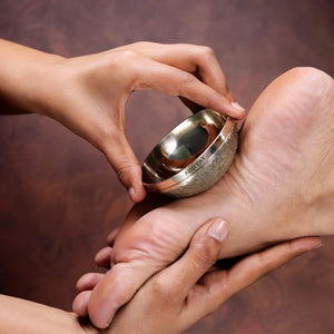 Hands using Adhyay Kansa Vatki bronze bowl to perform traditional foot massage therapy