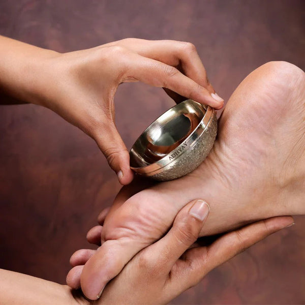 Hands using Adhyay Kansa Vatki bronze bowl to perform traditional foot massage therapy