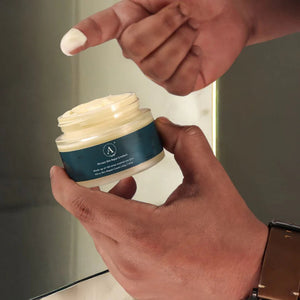 Man's hand applying Adhyay Nirvaan Skin Repair Cream made with 100 times washed cow ghee, holding open jar near mirror