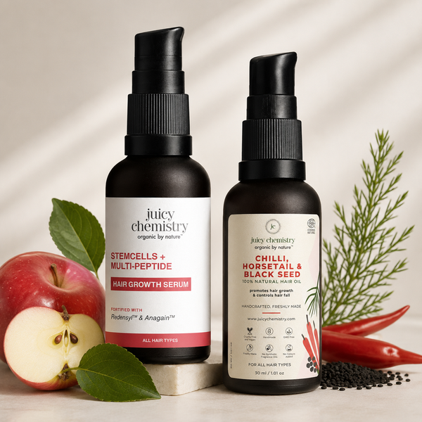 Two black pump bottles of Juicy Chemistry hair growth serums, one Stemcells + Multi-Peptide and one Chilli, Horserail & Black Seed, styled with fresh green leaves, a sliced red apple, whole and sliced red chilies, and black seeds on a light beige background.